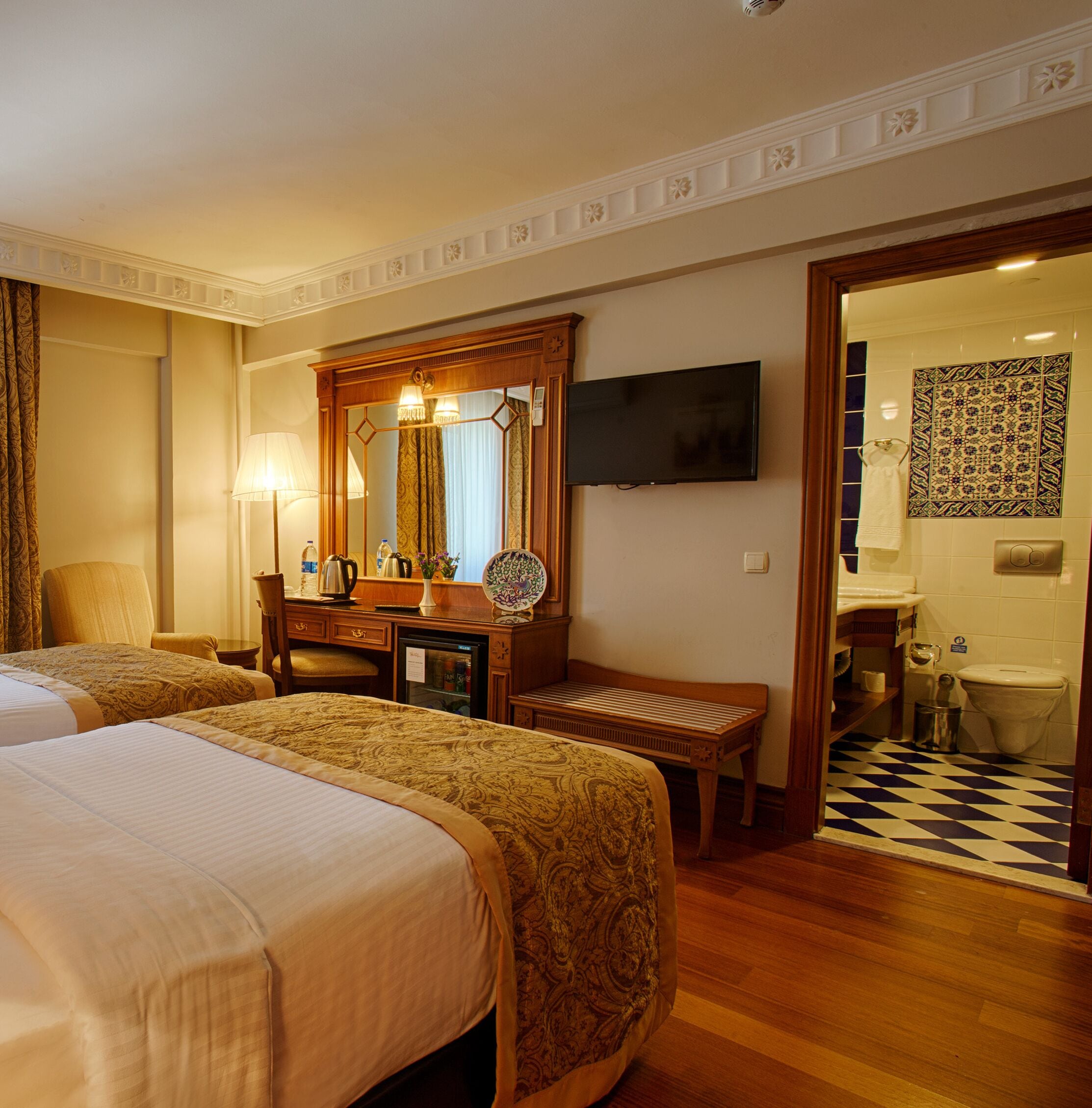 A hotel room with two beds, a desk with a mirror, a television, a bathroom with a checkered floor, and a patterned tile wall.