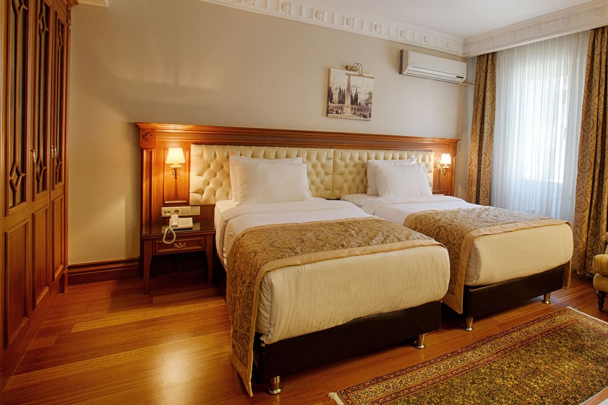 Habitación de hotel con dos camas, piso de madera, techo decorativo y una mesita de noche con lámpara.