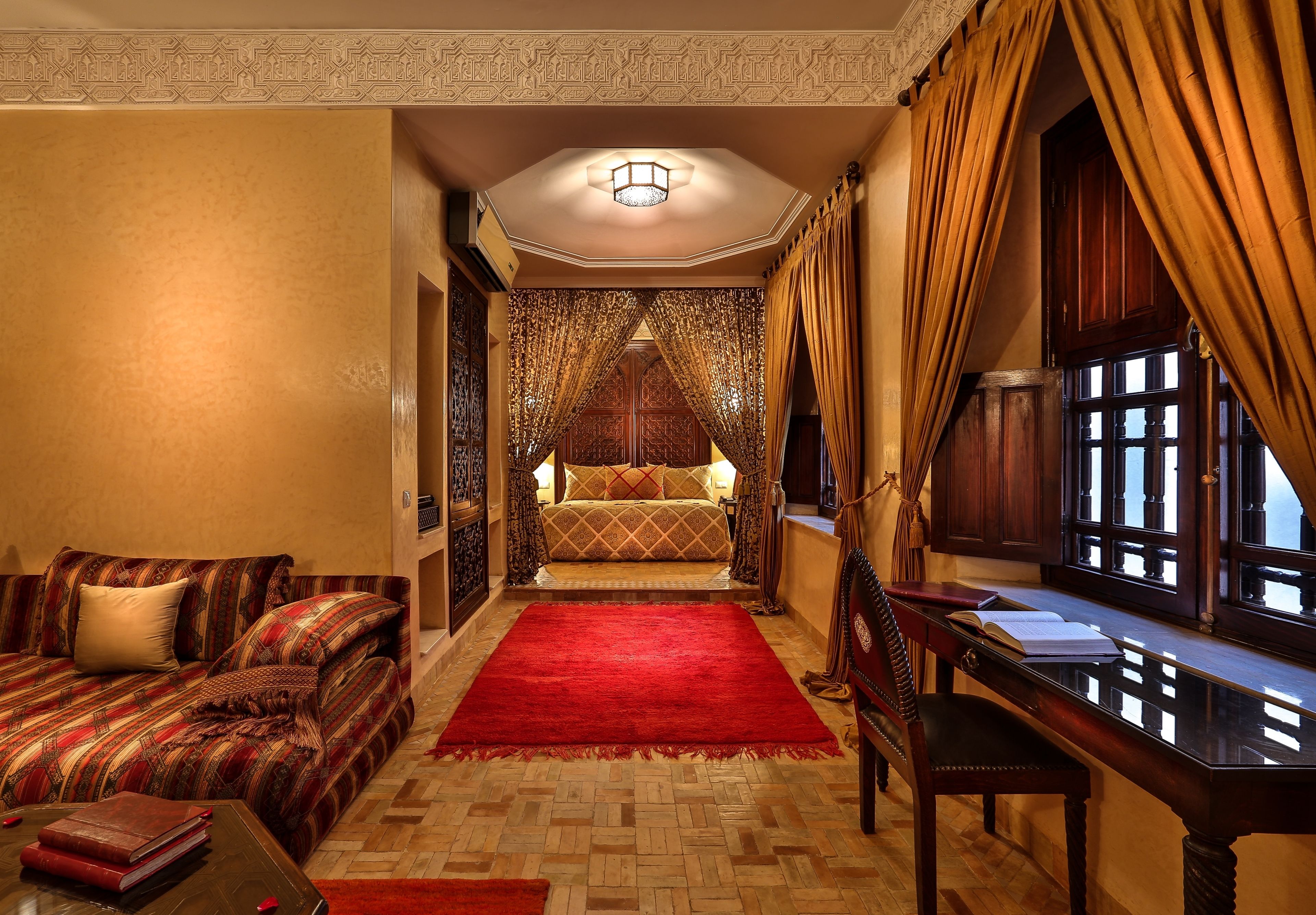 A room with a sofa, a red rug, a wooden desk, and a bed with patterned curtains.