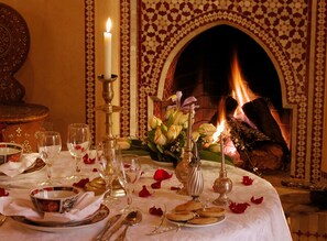 A dining table set with fine china, glasses, and a candle, with a fireplace in the background. - Riad Kniza (Marrakech)