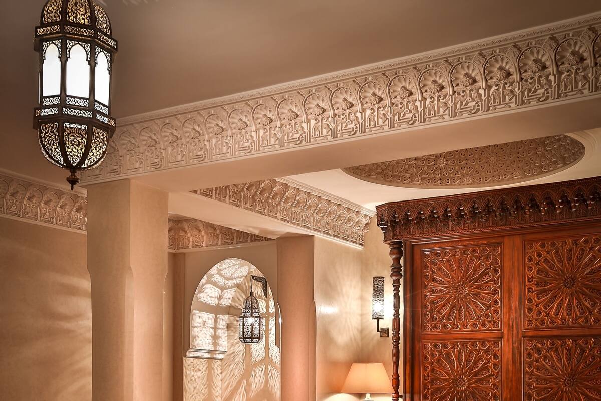 A traditional Moroccan interior with ornate wood paneling, a plush sofa set, a coffee table with fruit, and a chandelier.