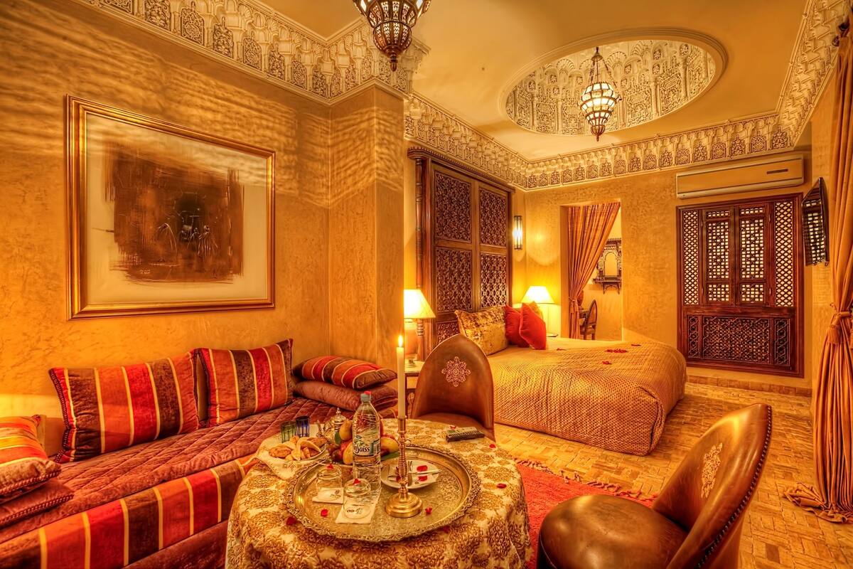 A luxurious hotel room with a large bed, a sofa, a dining table with a tray, and intricate wall and ceiling designs.