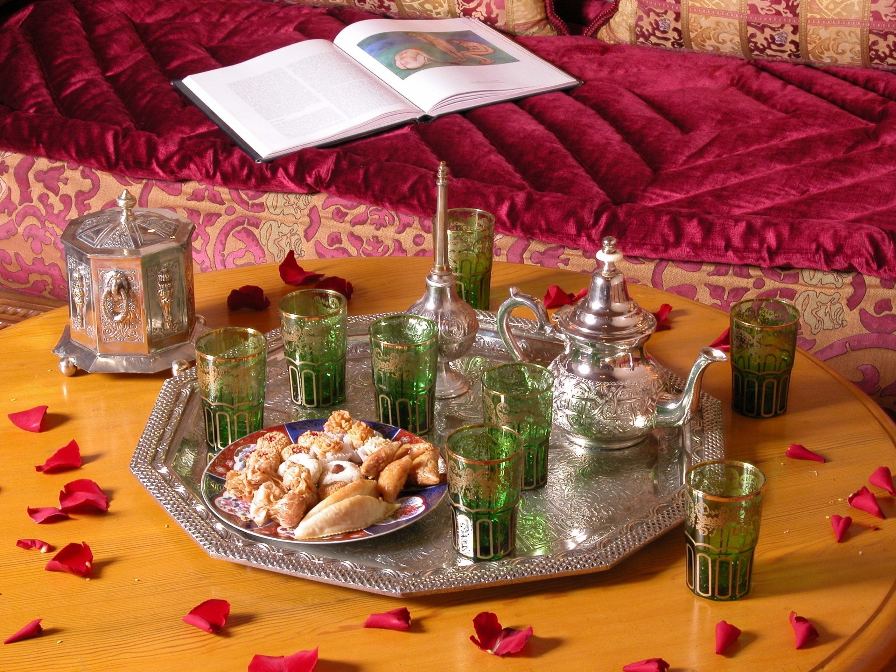 A tray with pastries, glasses, and a teapot on a wooden table with rose petals, set against a patterned sofa and a book.