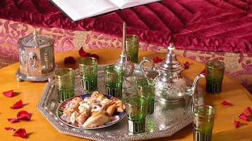 A tray with pastries, glasses, and a teapot on a wooden table with rose petals, set against a patterned sofa and a book.
