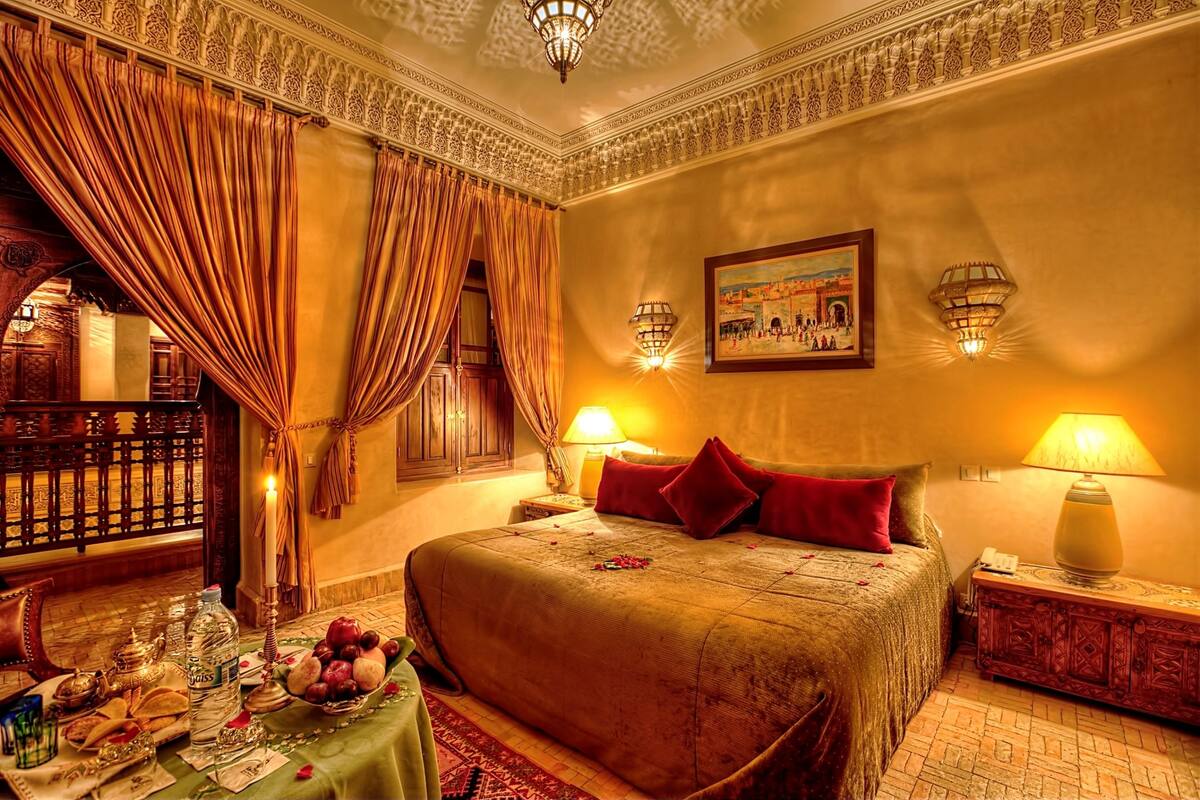 A luxurious hotel room with a large bed, ornate curtains, a chandelier, and a painting on the wall.