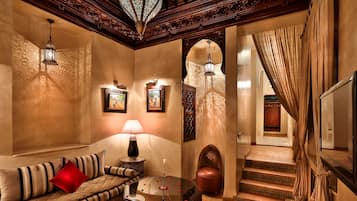 A cozy living room with a sofa, a coffee table, and a lamp. There are framed pictures on the wall and a staircase leading to another room.