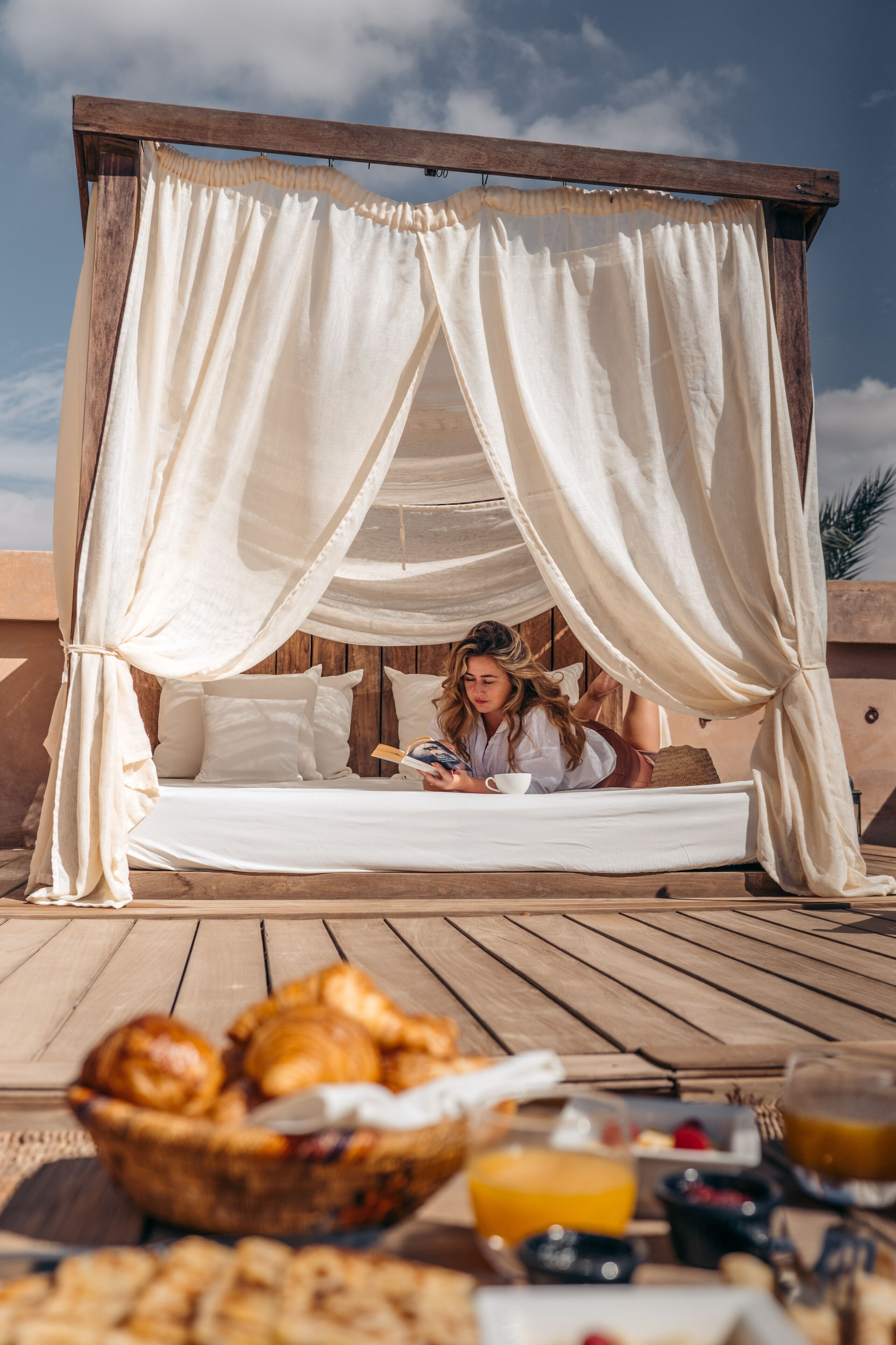 Un lit à baldaquin, un plateau de petit-déjeuner avec des viennoiseries et des boissons, et une terrasse en bois.