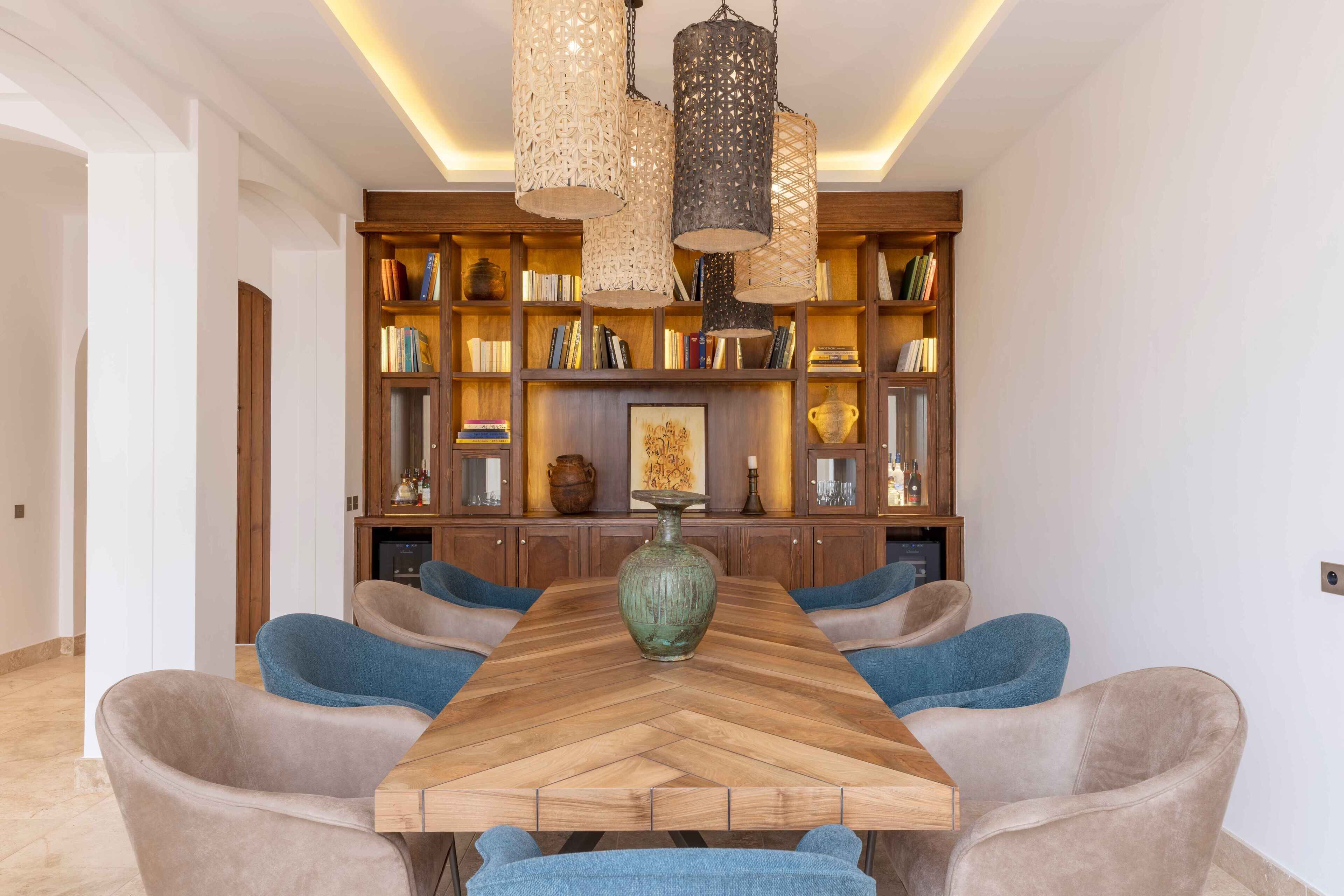 Un'area pranzo con un lungo tavolo in legno, sedie rivestite di blu e una libreria piena di libri e oggetti decorativi.