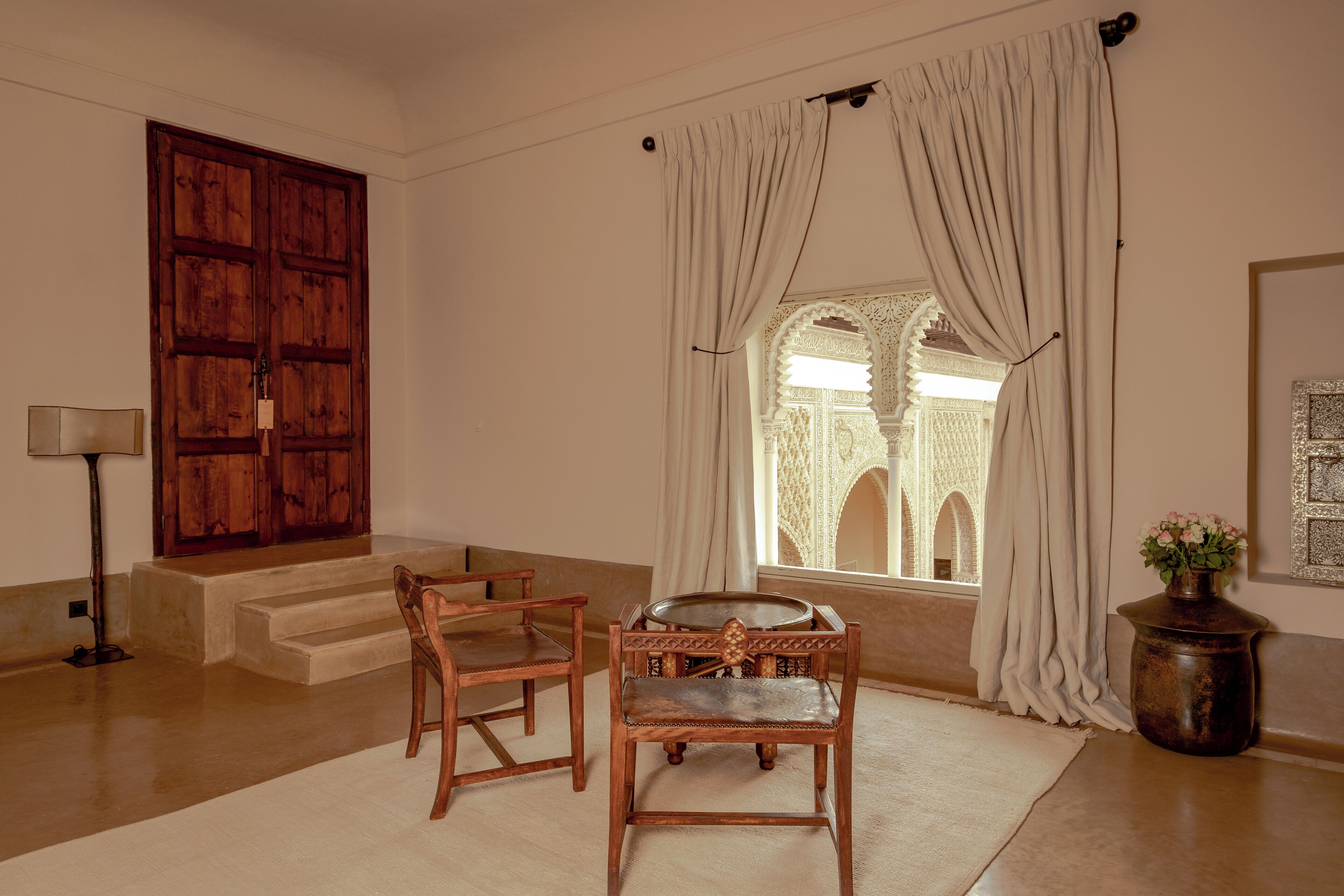 Una habitación con puerta de madera, ventana con cortinas, una mesita, dos sillas, una lámpara y un jarrón con flores.