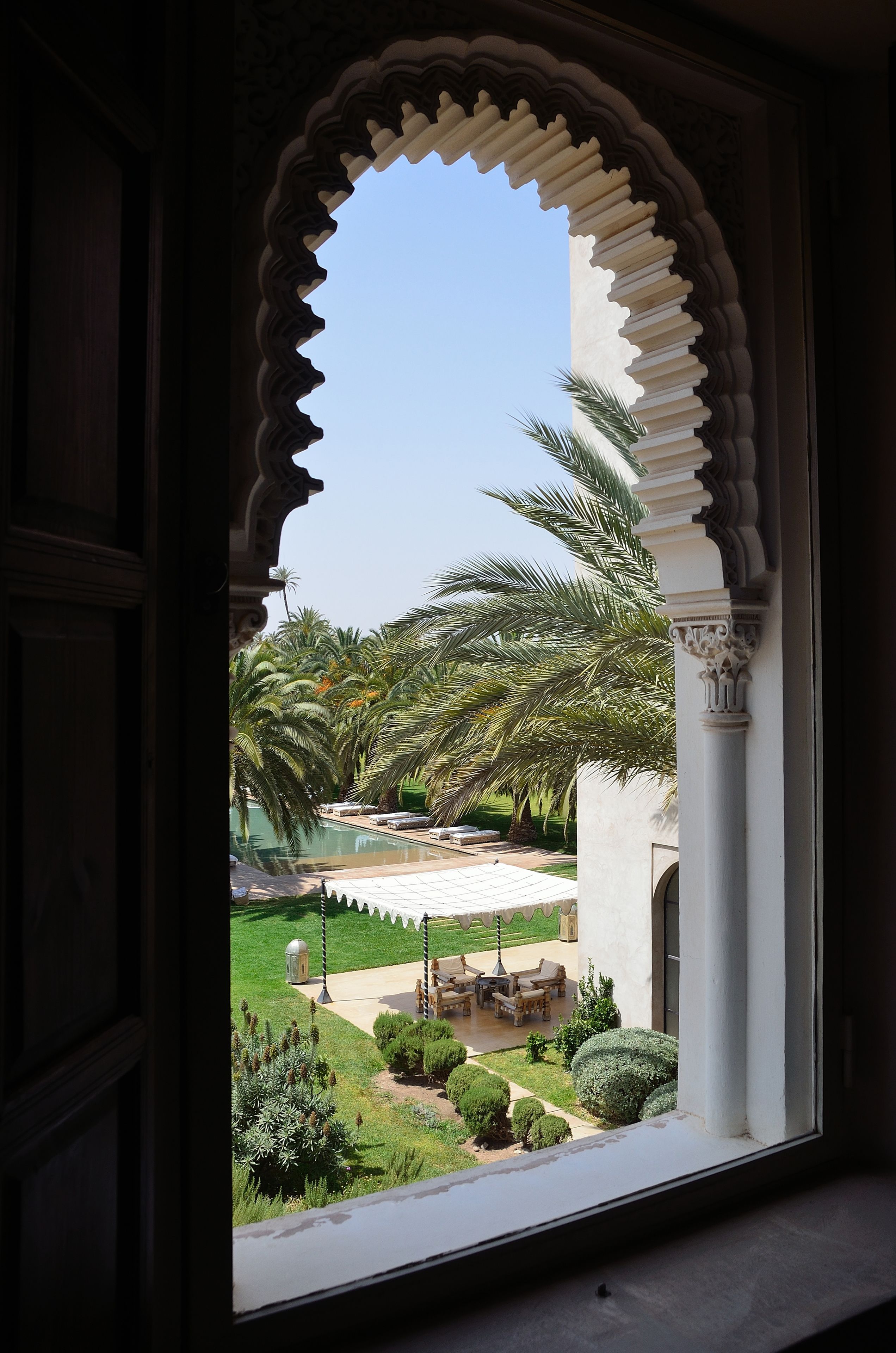 A view through an ornate doorway to a courtyard with palm trees, a pool, and outdoor furniture.