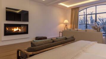 A modern bedroom with a fireplace, a large bed, a sofa, and a view of palm trees through a window.