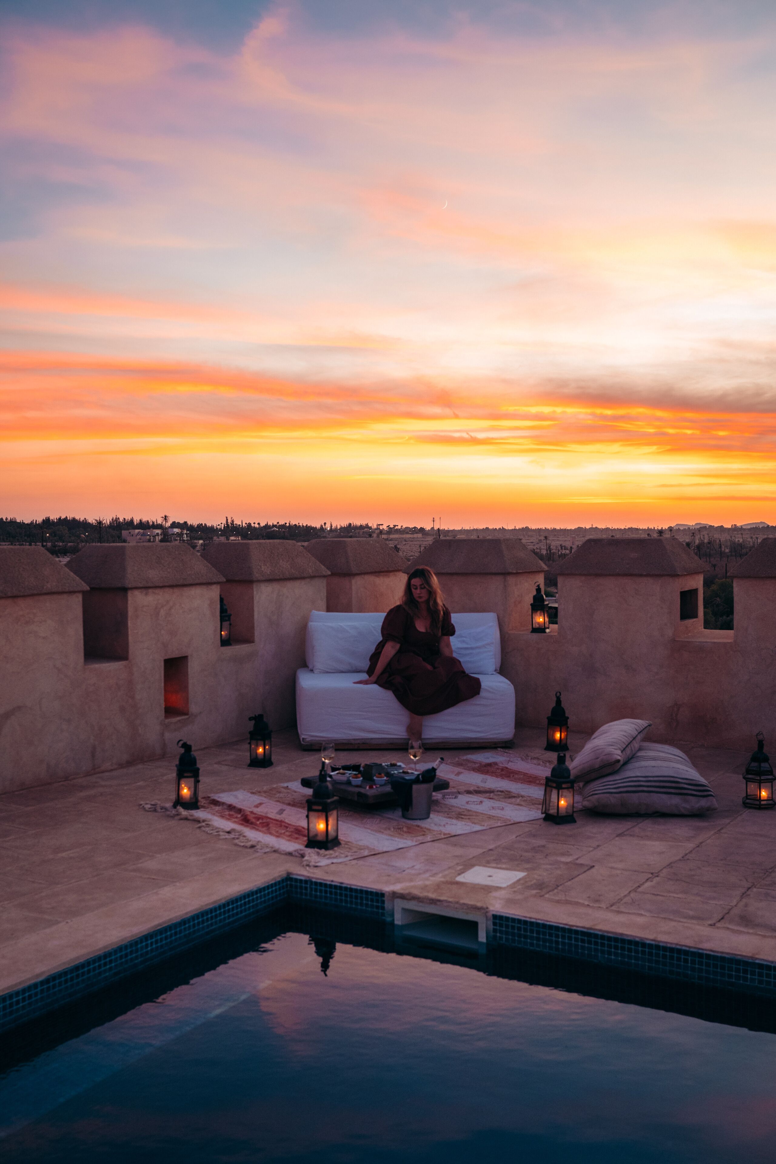 Un área de piscina en la azotea con una cama y sillas de descanso, rodeada de faroles, durante el atardecer.