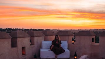A rooftop pool area with a bed and lounge chairs, surrounded by lanterns, during sunset.