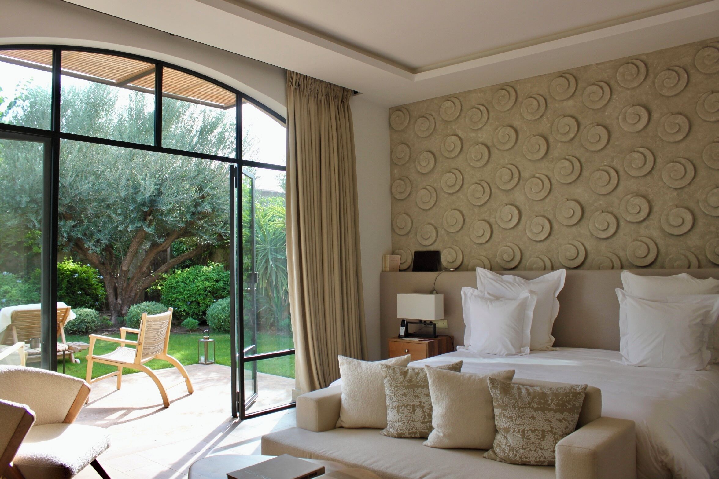 Un dormitorio con una cama grande, un sofá y una silla. Se ve vegetación a través de la ventana.