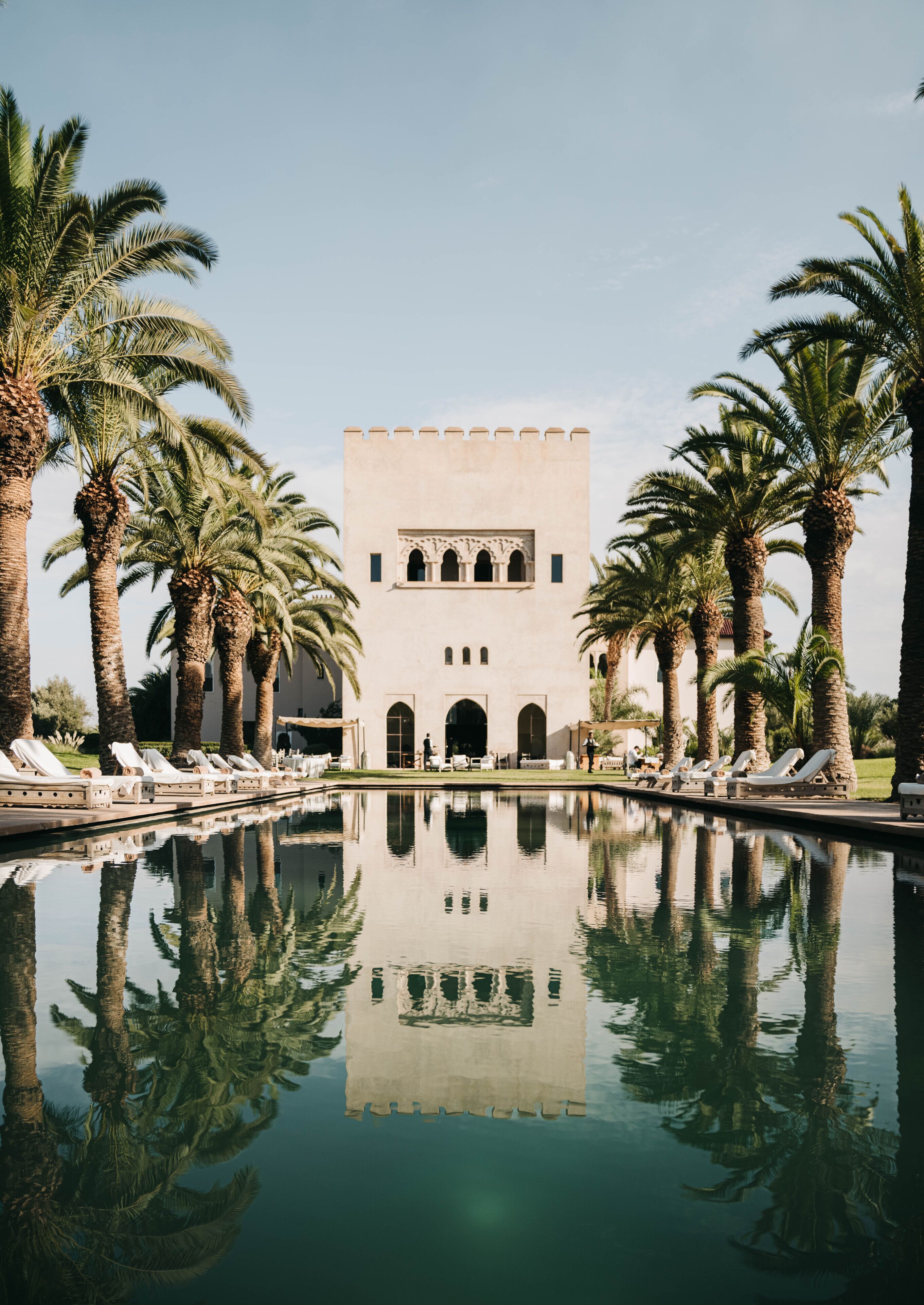 Ein Pool mit Palmen und ein Gebäude mit Rundbogenfenstern und einem Turm.