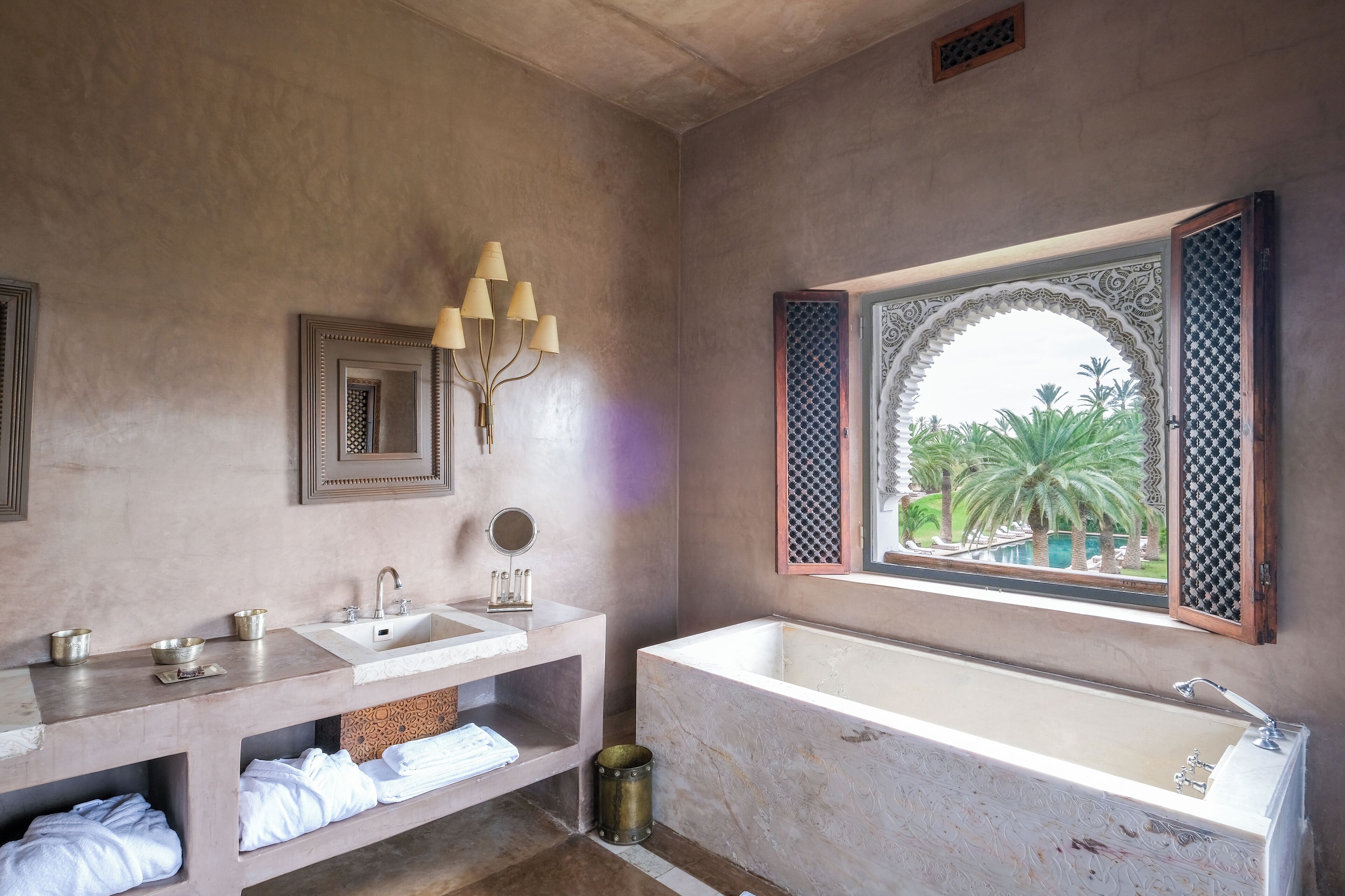 A bathroom with a large bathtub, a sink, and a mirror. There is a window with a decorative frame, and towels are neatly arranged on shelves.