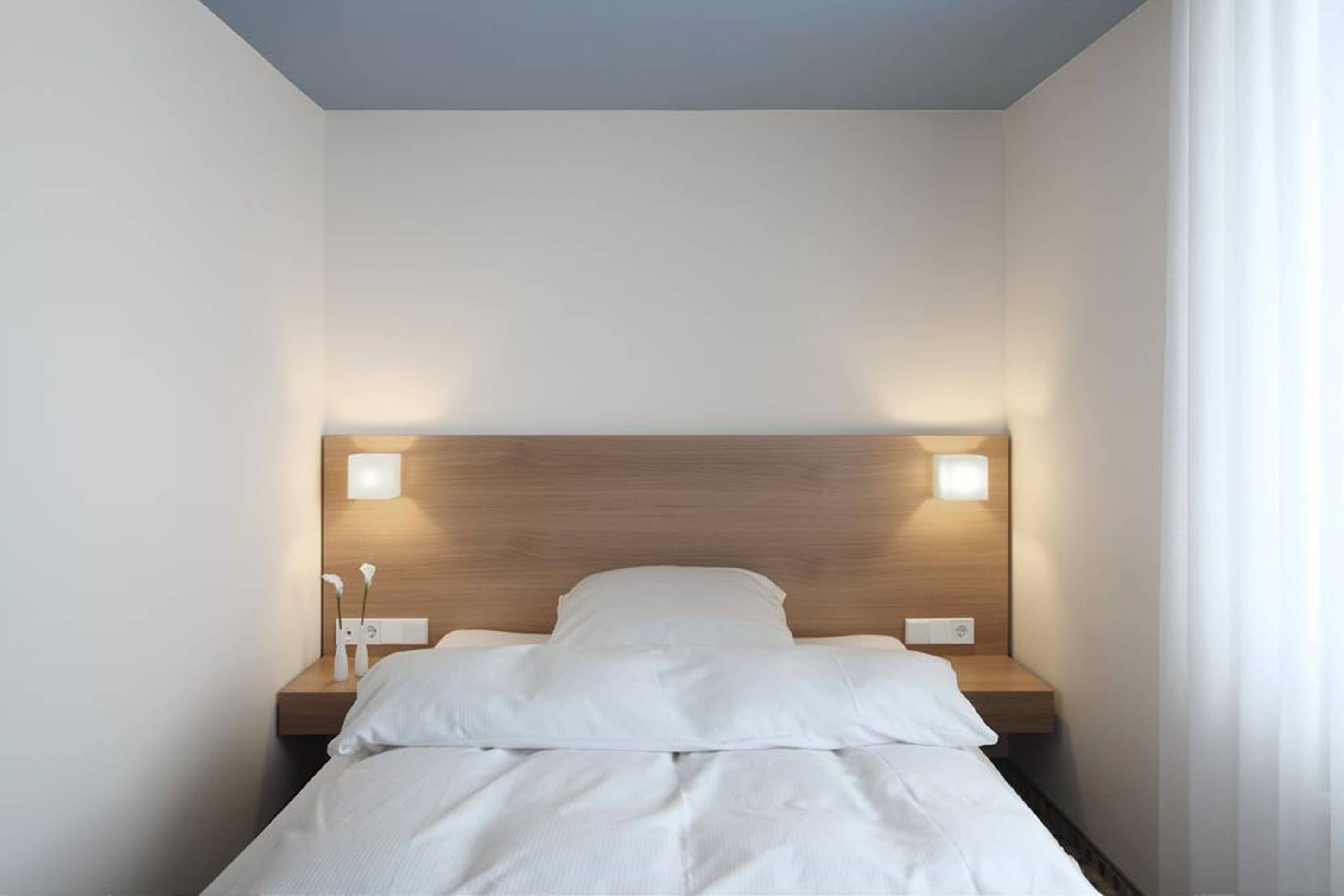 Uma cama bem arrumada com lençóis brancos, uma cabeceira de madeira e duas luminárias laterais.