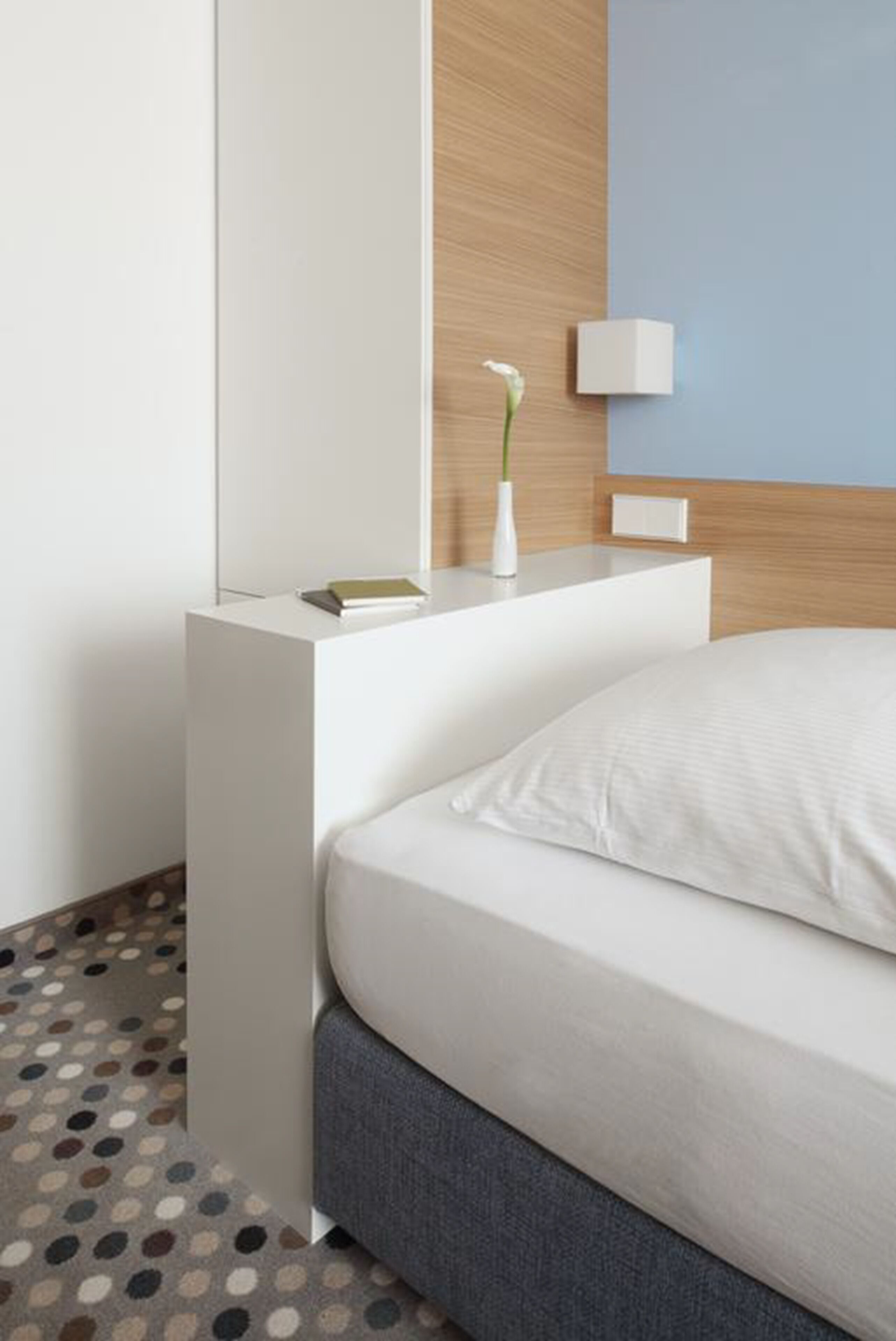A modern hotel room with a neatly made bed, a bedside table with a book and a vase, and a wall-mounted light.