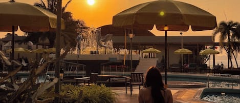 Une vue au coucher du soleil, avec une femme assise à une table, des parasols et un espace piscine.