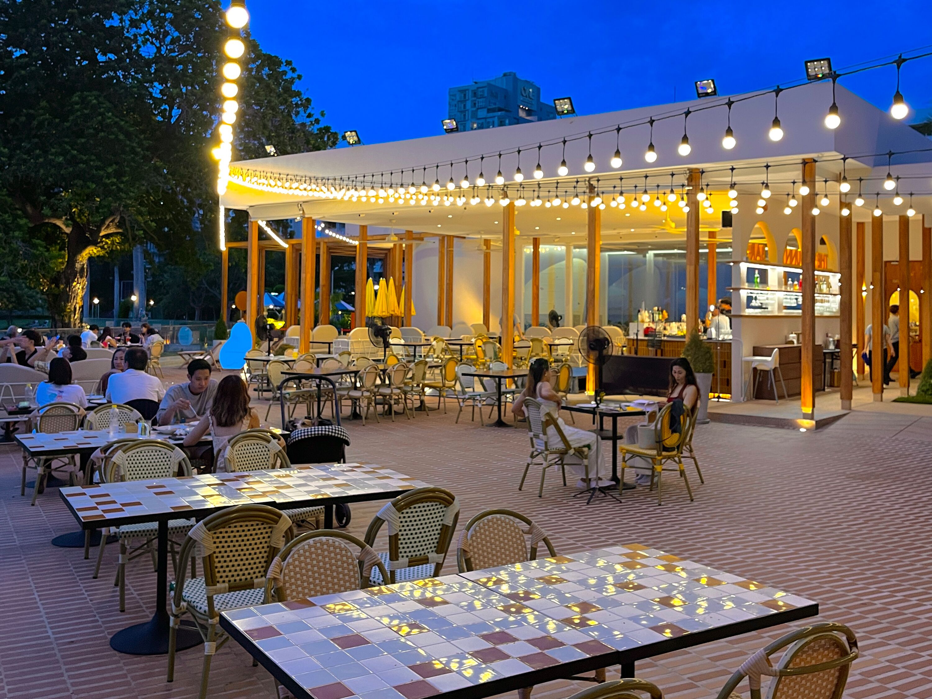 An outdoor dining area with tables and chairs, a covered section with a bar, and a view of the cityscape in the background.