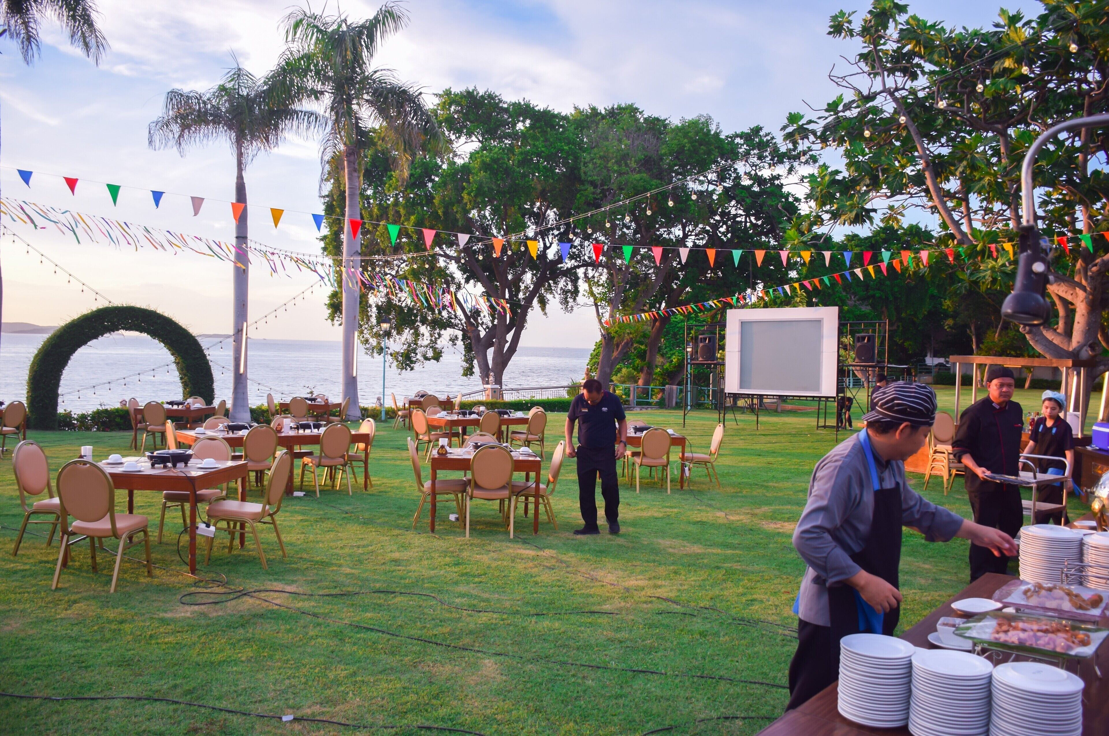 An outdoor dining area with tables and chairs, a buffet setup, and a large screen in the background.