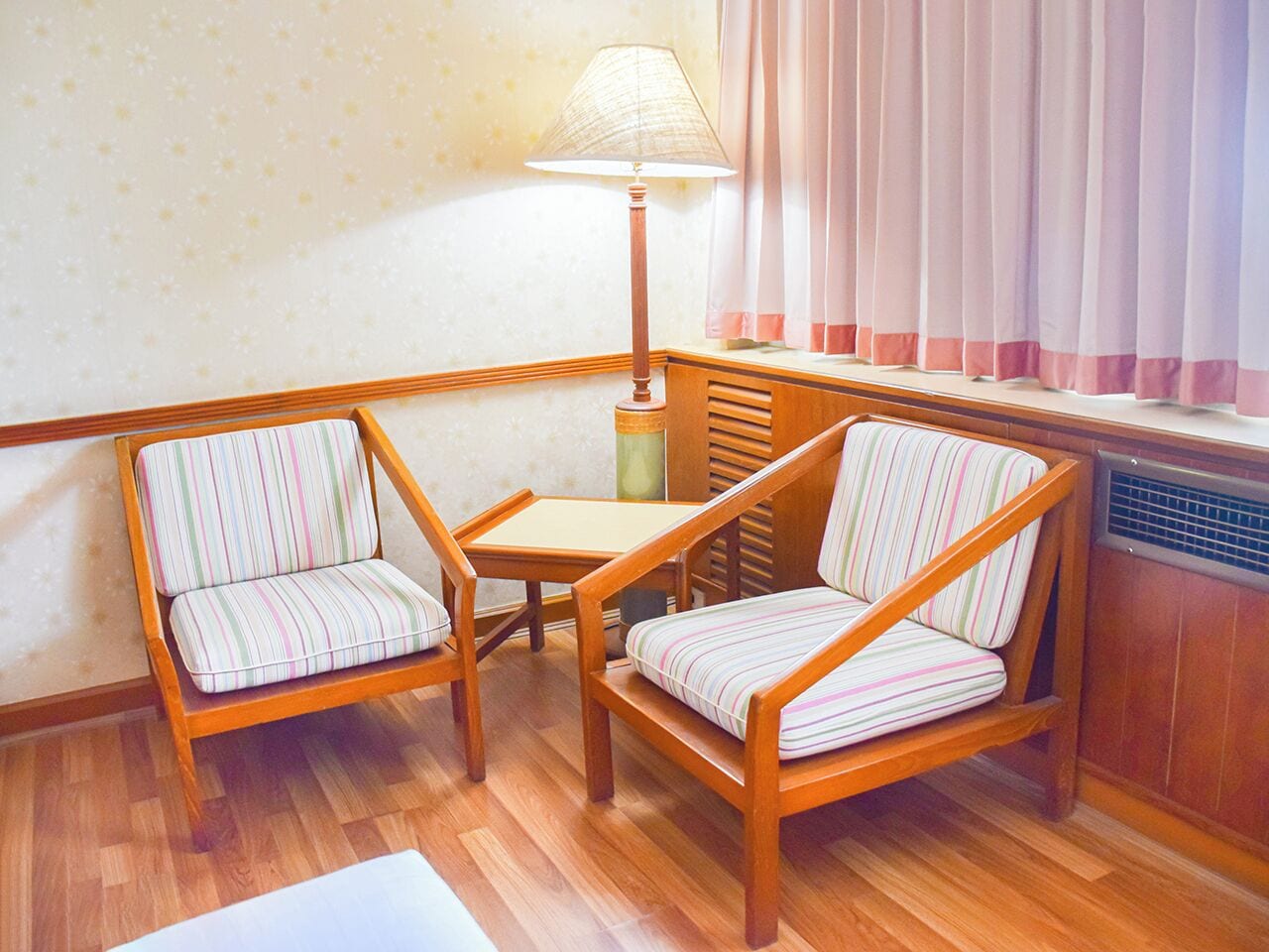 A corner room with two striped armchairs, a small wooden table, a floor lamp, and a radiator.