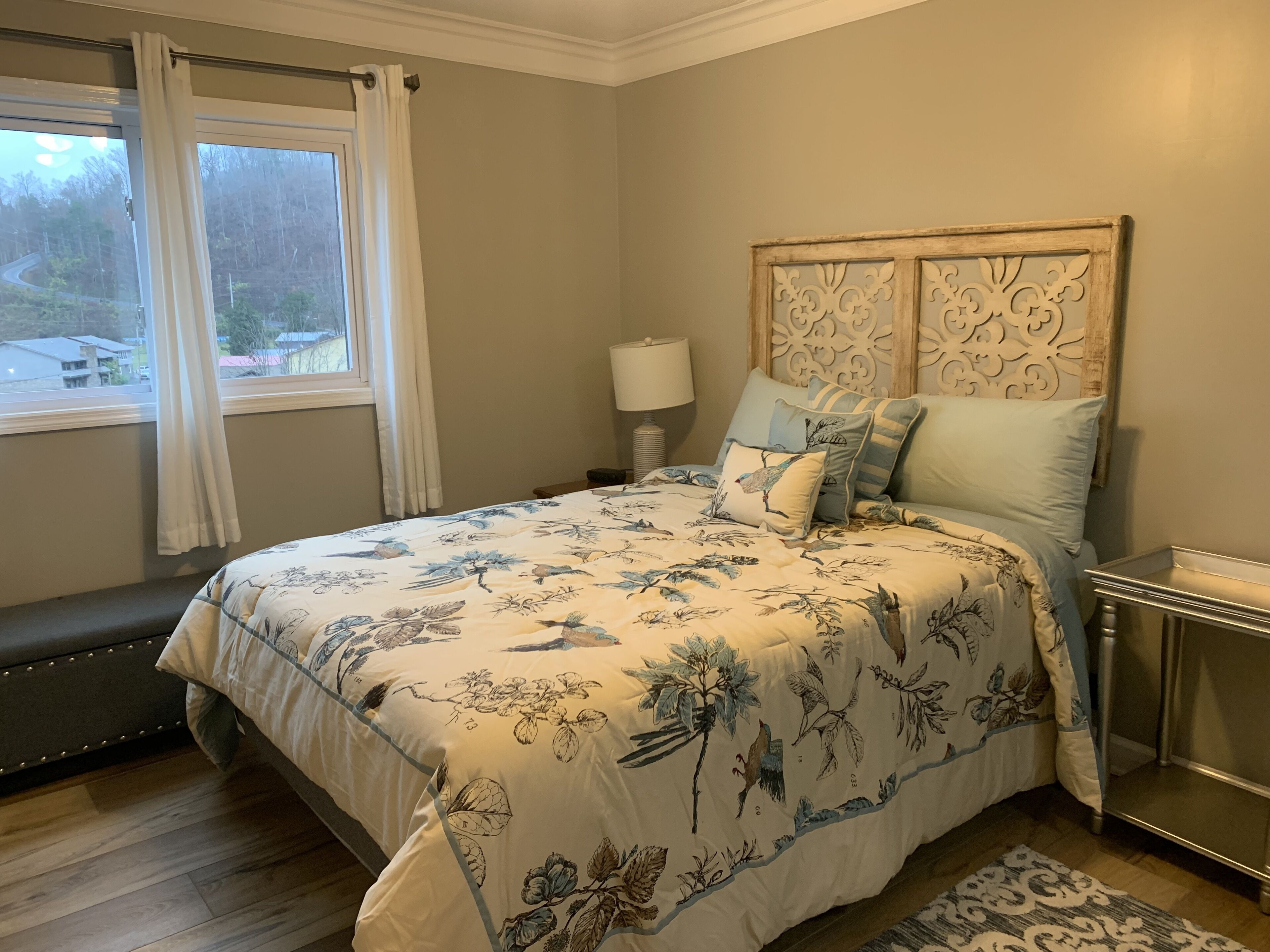 A bedroom with a large bed, a nightstand, a lamp, and a view of a forest through a window.