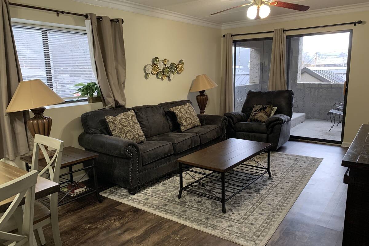A living room with a dark brown leather sofa, two armchairs, a coffee table, and a dining table with chairs. There are two lamps, a decorative wall piece, and a view through a sliding glass door.