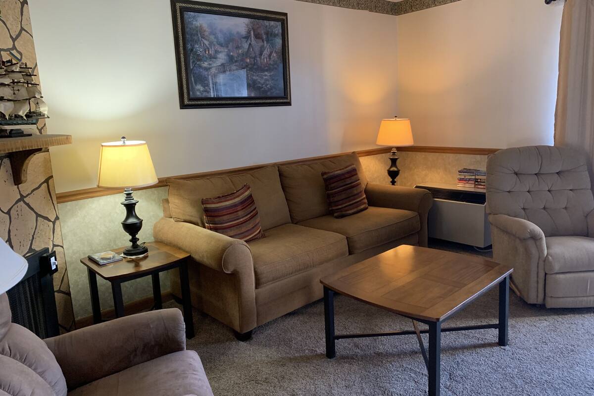 A cozy living room with a sofa, armchairs, a coffee table, and a fireplace. There is a painting on the wall and a lamp on the side table.