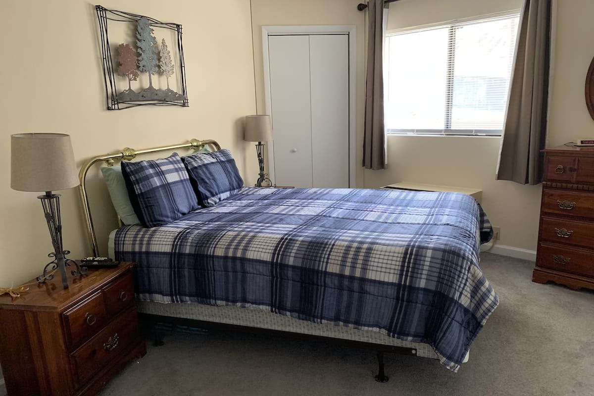 A neatly made bed with a plaid bedspread, a bedside table with a lamp, a dresser, a door, and a window with curtains.