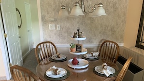 A dining area with a round wooden table, four chairs, a chandelier, and a three-tiered serving tray with food and a sign.
