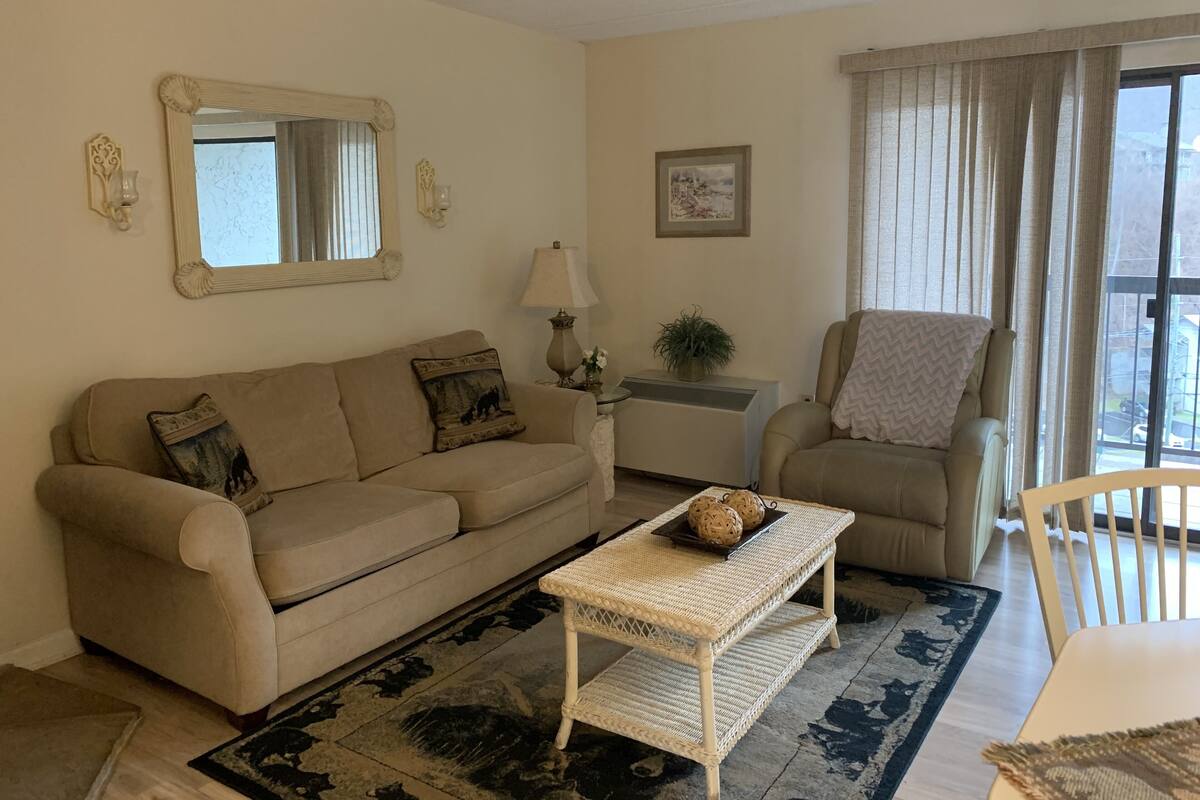 A living room with a beige sofa, a white coffee table, a chair, a lamp, and a large window with curtains.