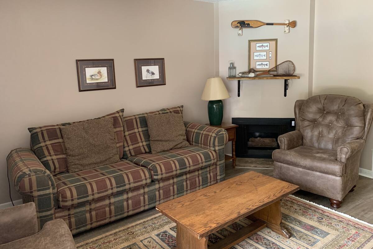 A living room with a plaid sofa, a leather armchair, a wooden coffee table, a fireplace, and framed pictures on the wall.