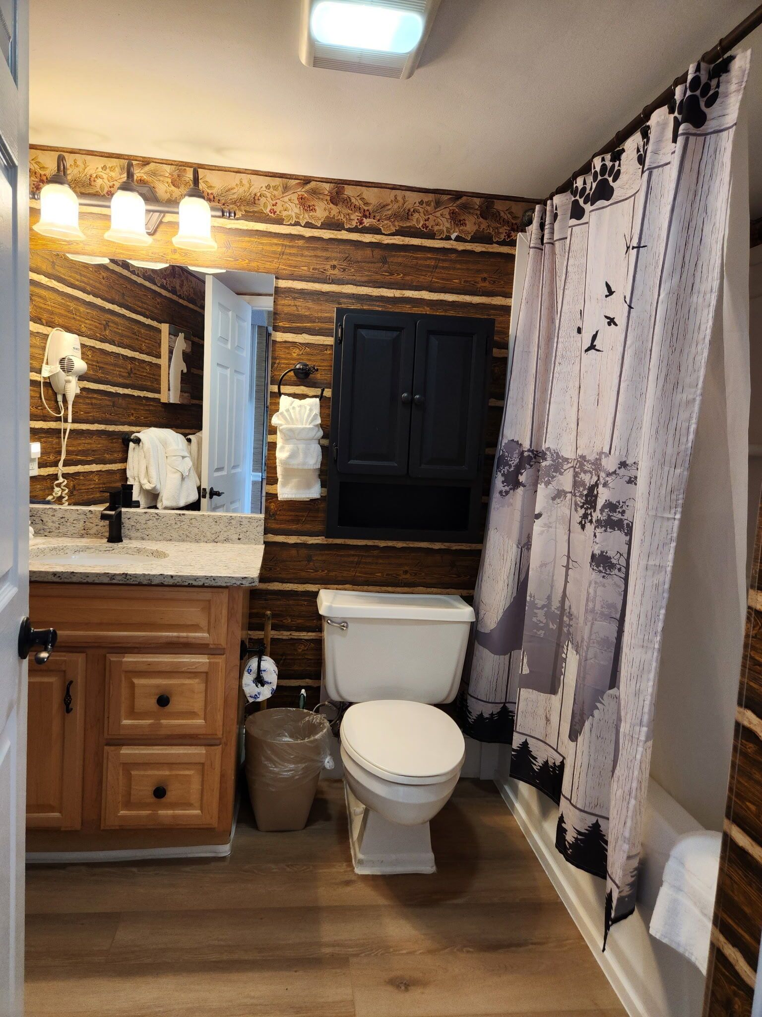 Ein Badezimmer mit einer Holzwand, einer weißen Toilette, einem Waschbecken mit Spiegel und einer Duschvorhang mit Waldmusterung.