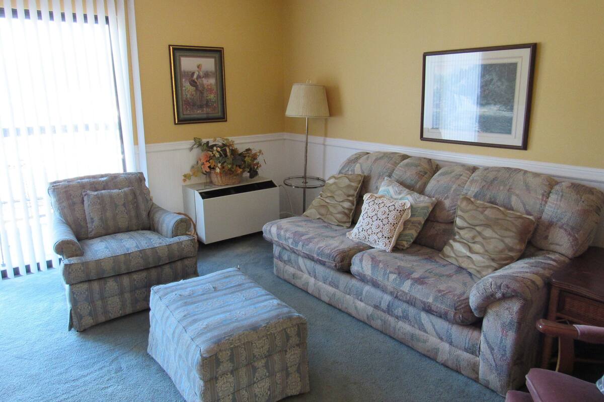 A living room with a sofa, armchair, ottoman, and a side table with a lamp.