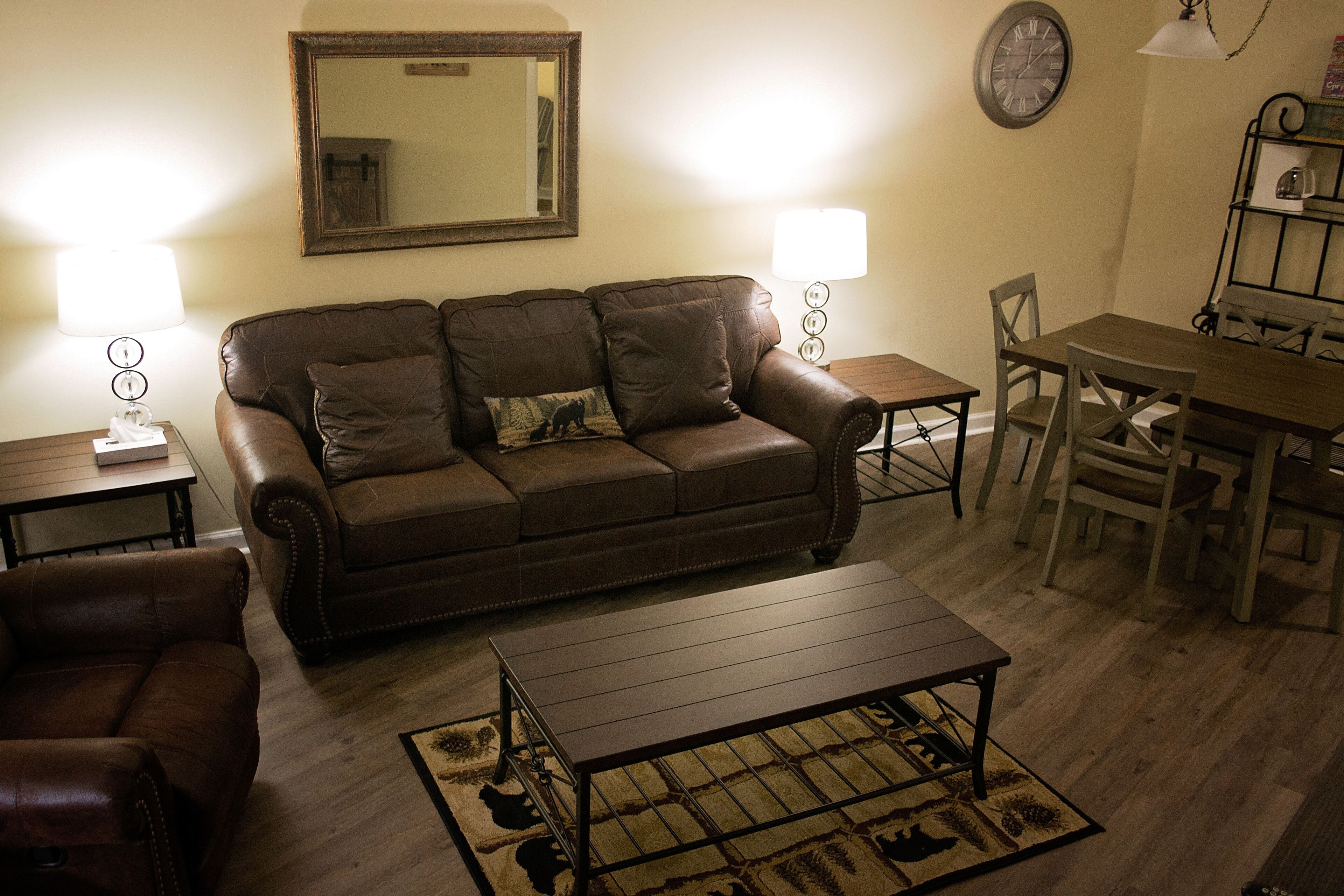 A living room with a brown leather sofa, a wooden coffee table, and a dining table with chairs. There are two lamps on either side of the sofa and a clock on the wall.