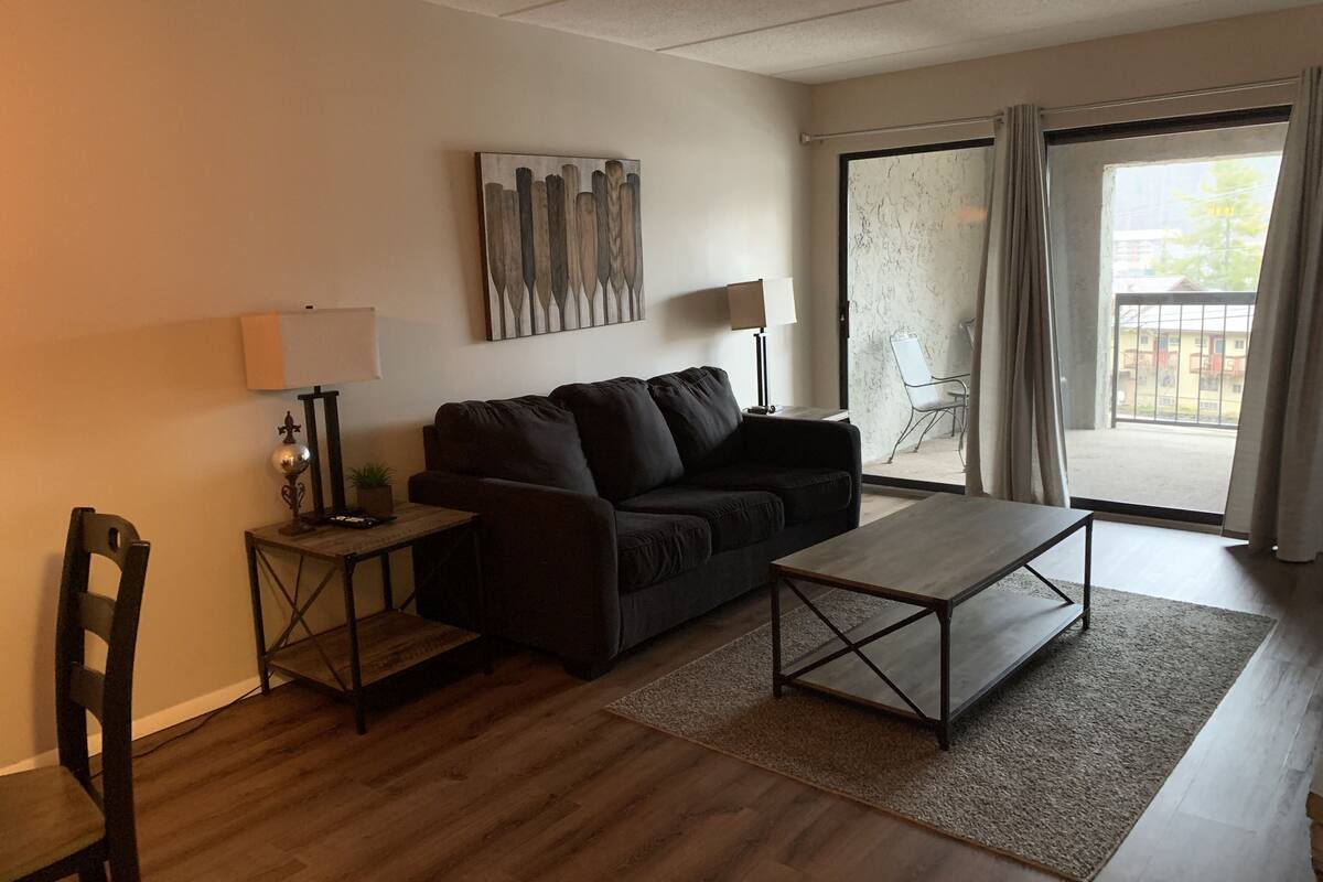 A living room with a sofa, a coffee table, a lamp, and a chair. There is a window with curtains and a view of buildings outside.