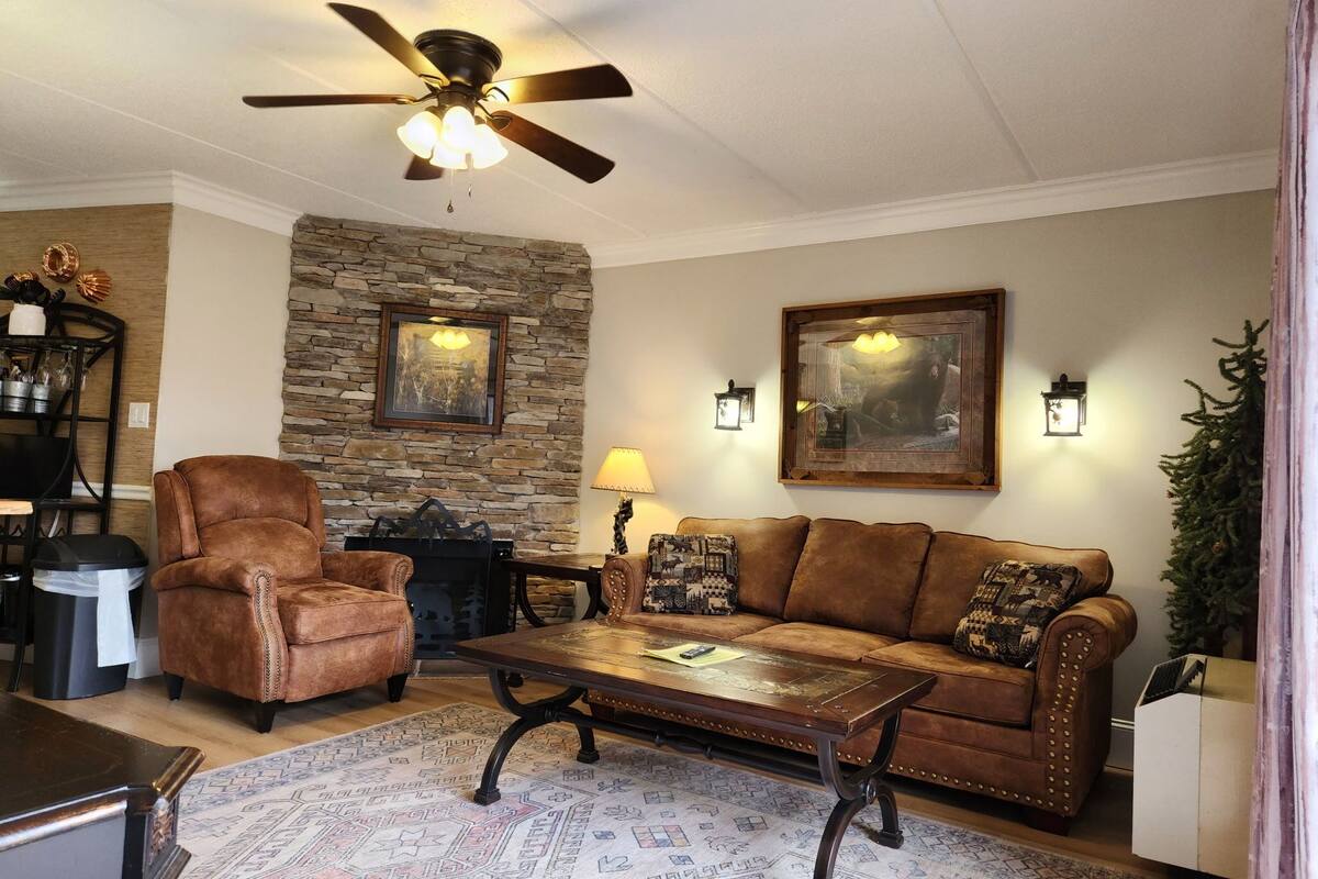 A living room with a stone wall, a ceiling fan, a brown leather armchair, a brown sofa, a wooden coffee table, and patterned rugs.