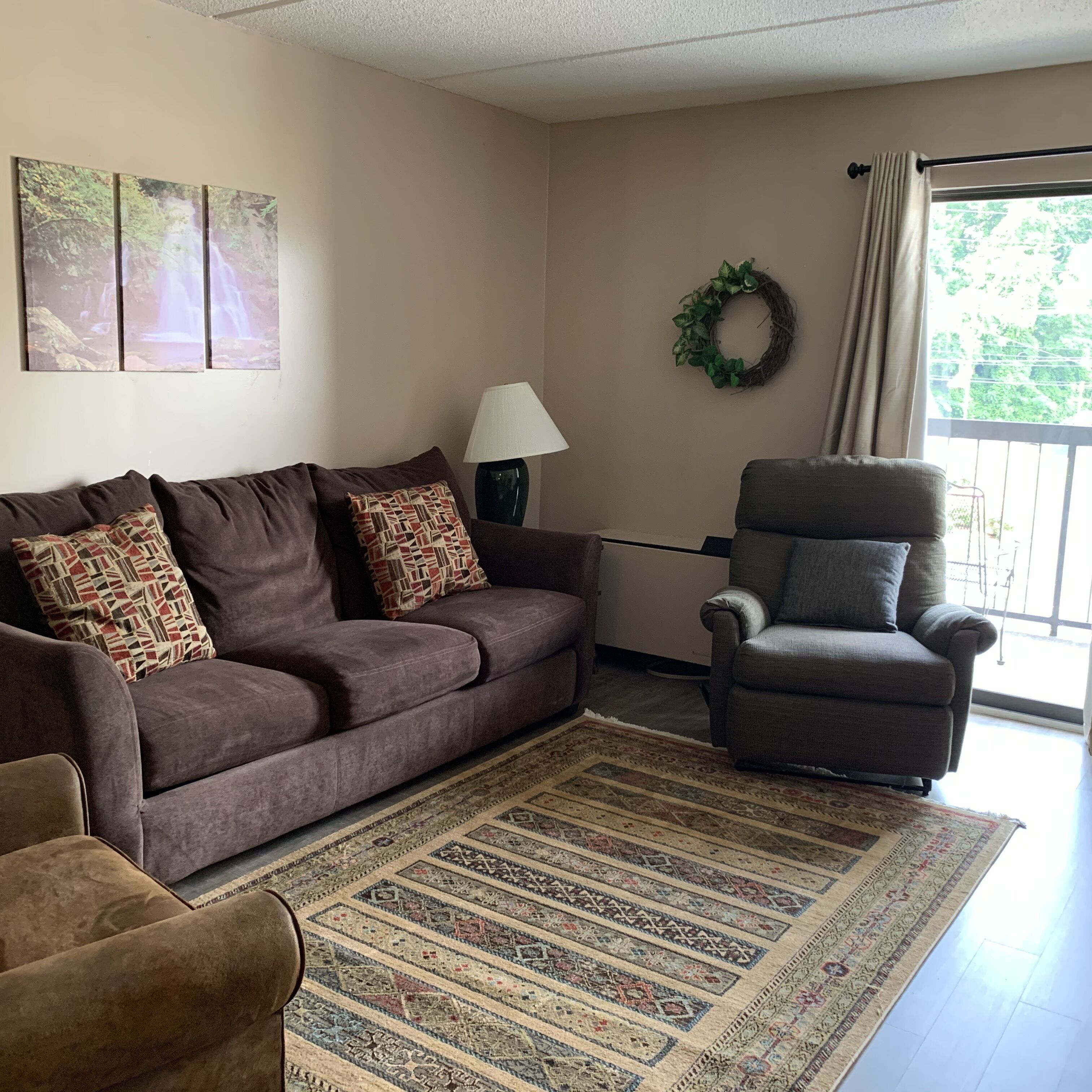 A living room with a brown sofa, patterned cushions, a grey armchair, a floor lamp, a decorative wreath, and a large window with curtains.