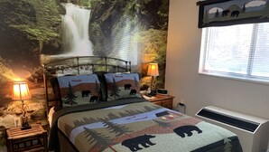 A bedroom with a bed featuring a forest-themed quilt, two bedside lamps, a nightstand, and a wall-mounted TV displaying a bear illustration.