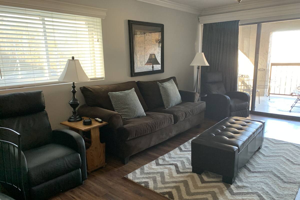 A living room with a brown sofa, a black armchair, a wooden side table, a lamp, and a patterned rug.