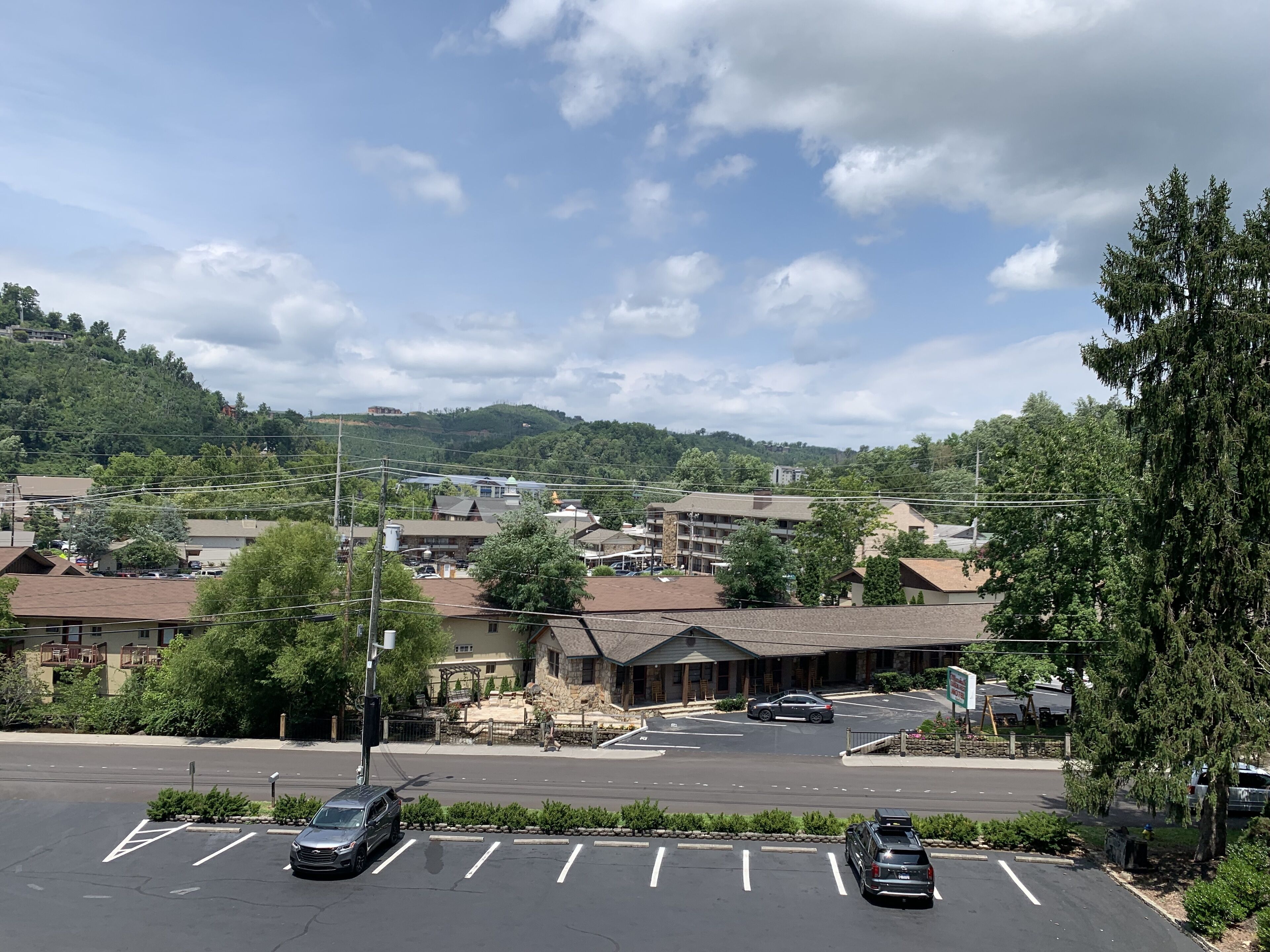A parking lot with multiple cars, a building with a stone facade, and a hilly landscape in the background.