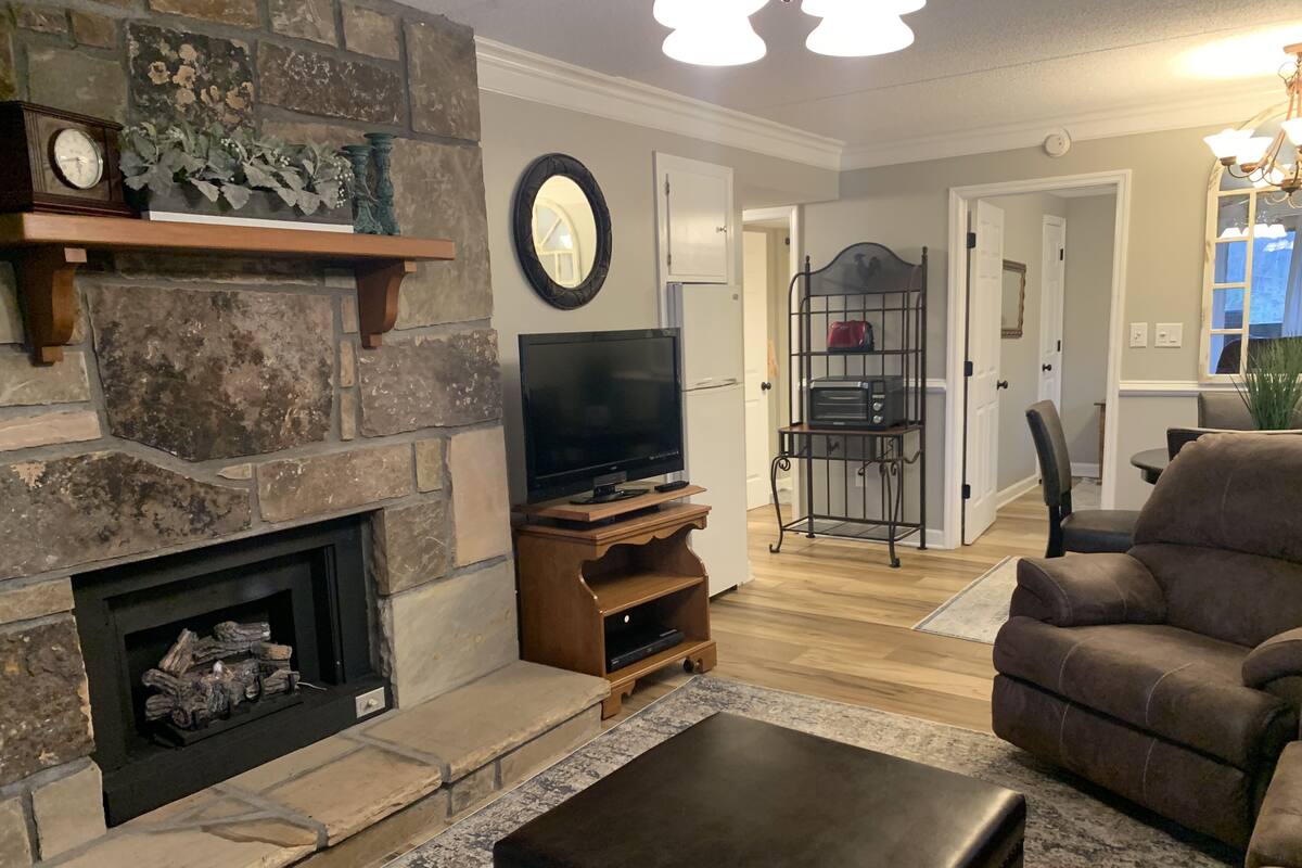 A living room with a stone fireplace, a flat-screen TV, and a brown leather ottoman.