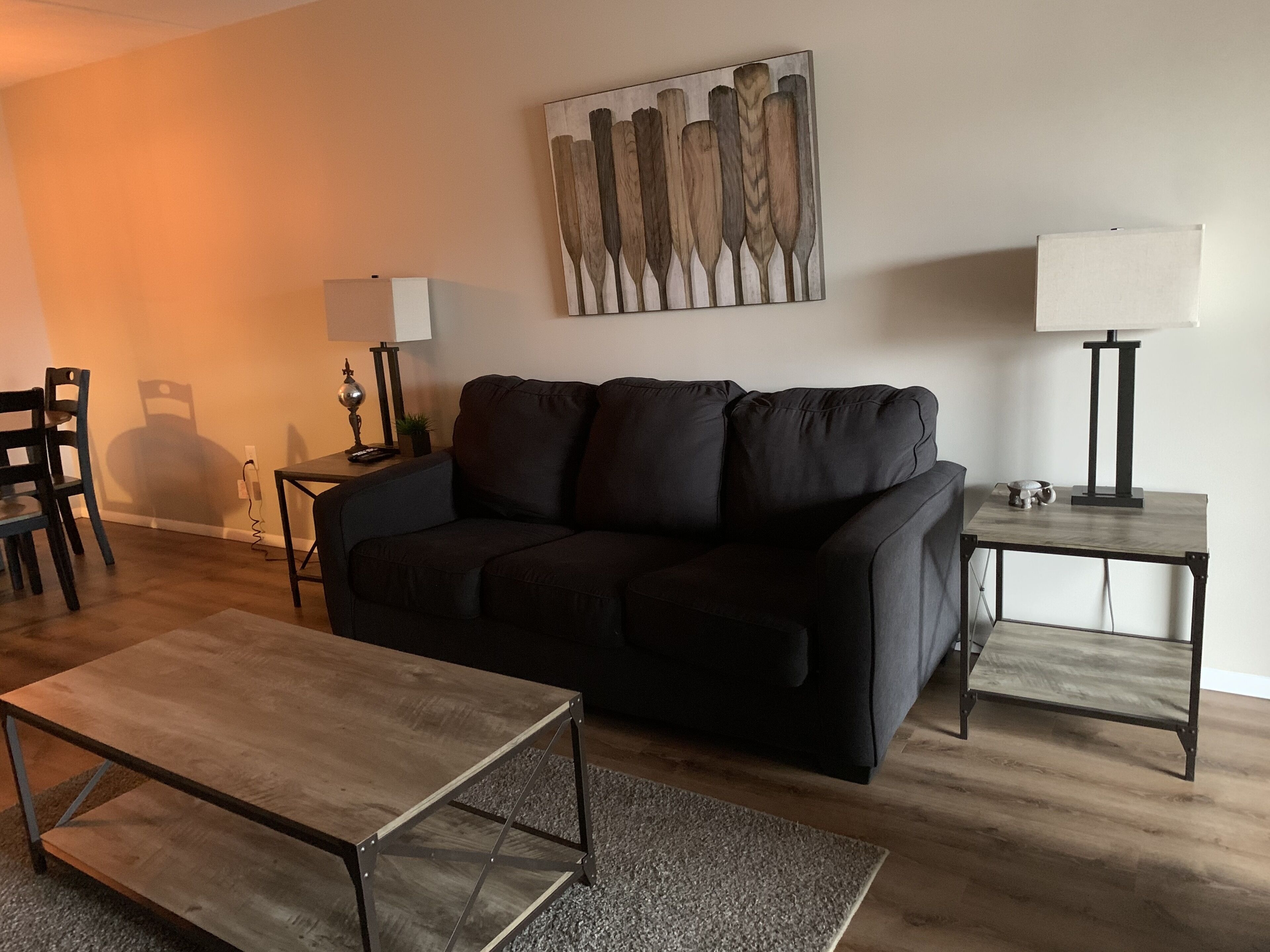 Una sala moderna con un sofá negro, una mesa de centro de madera, un cuadro en la pared y una lámpara sobre una mesita auxiliar.