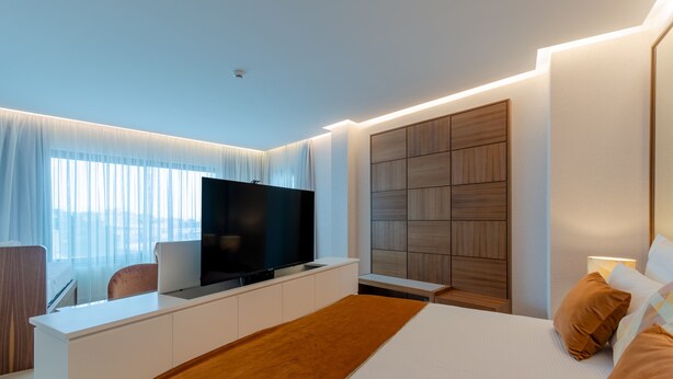 A modern hotel room with a large bed, a flat-screen TV, a desk with a laptop, and a large window with sheer curtains. - Hotel Alvorada (Cascais)