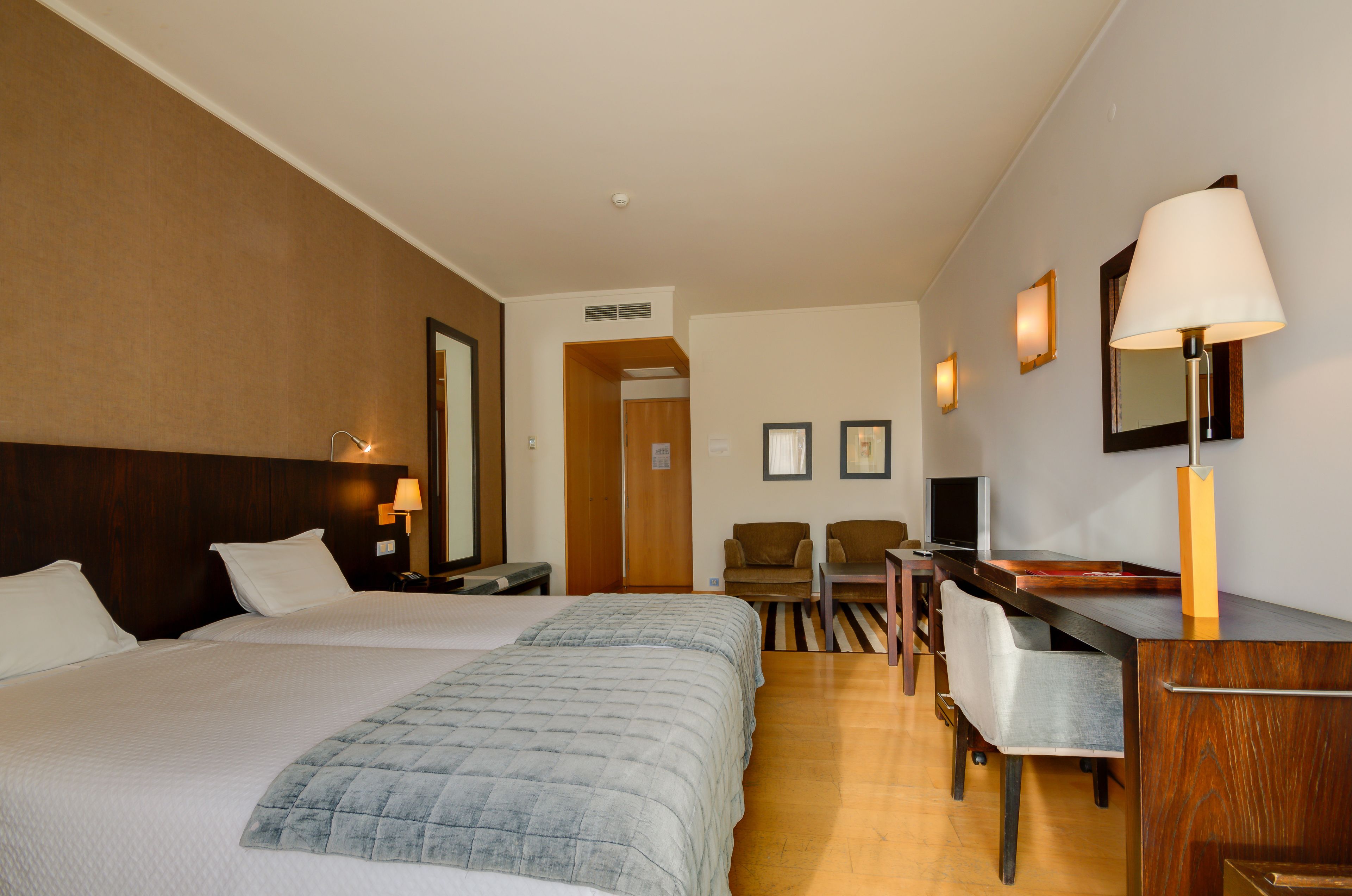 A hotel room with two beds, a desk, a chair, a TV, and a lamp.