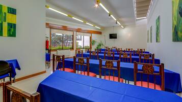 Un salón de conferencias con mesas y sillas azules, ventanas amplias y un televisor de pantalla plana en la pared.