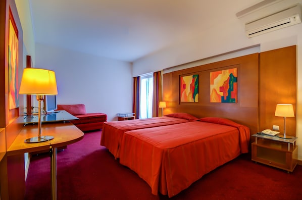 A hotel room with two beds, a desk with a lamp, a chair, a small table, and artwork on the wall.