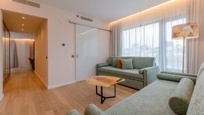 A modern living room with a sofa, a coffee table, and a view of the city through large windows. - Hotel Alvorada (Cascais)