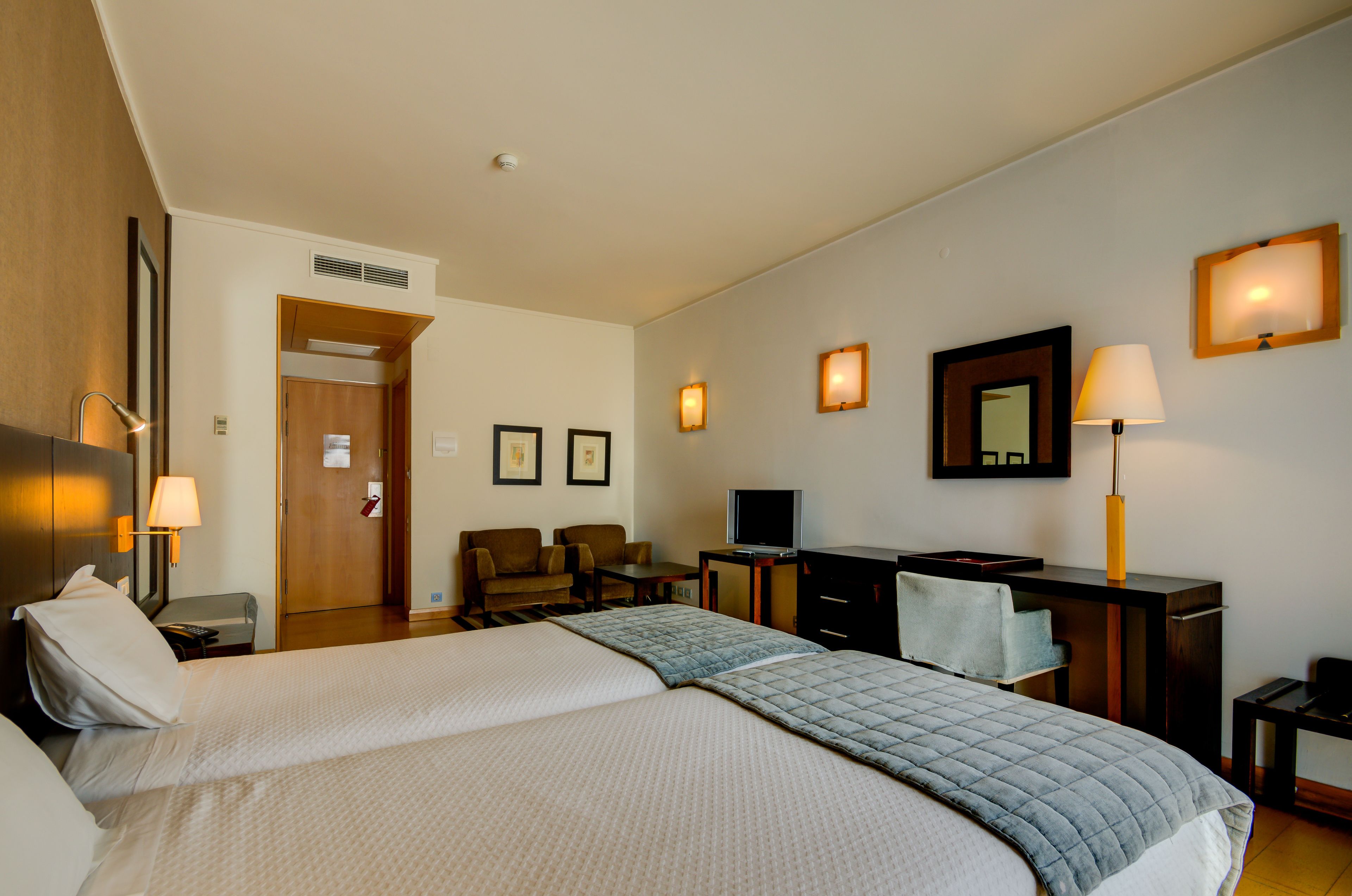 A hotel room with two beds, a desk, a chair, a TV, and a door leading to another room.